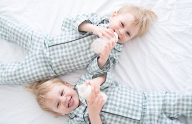 Pijamalı iki bebek ikiz çocuk şişelerden süt içerek yatağa uzanıyorlar. Samimi bir çocukluk hayatı.