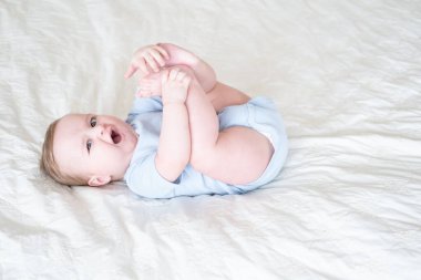 funny 6 months baby boy playing with legs, lying on bed at home on white bedding