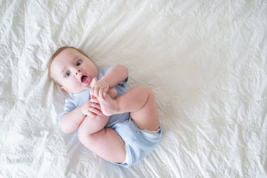 funny 6 months baby boy playing with legs, lying on bed at home on white bedding. top view