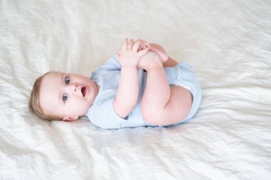 funny 6 months baby boy playing with legs, lying on bed at home on white bedding