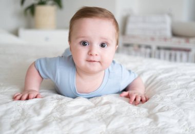 6 aylık mavi elbiseli komik bir bebek. Gülümsüyor ve evde yatakta yatıyor.