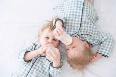 Pijamalı iki bebek ikiz çocuk şişelerden süt içerek yatağa uzanıyorlar. Samimi bir çocukluk hayatı.