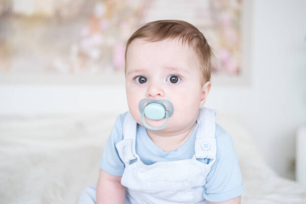 close up portrait baby boy 6 months old with nipple in blu clothes and sitting on white bed at home