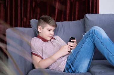 Genç yakışıklı çocuk akıllı telefondan medya içeriğini izliyor. Evdeki gri kanepede oturuyor.