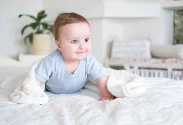 Симпатичный мальчик 6 месяцев в костюме blu body улыбается и лежит дома на кровати с белой кожей. — стоковое фото