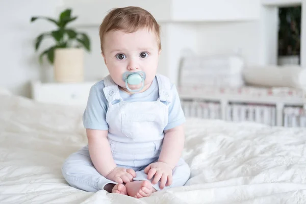 Baby boy 6 months old with nipple in blu clothes and sitting on white bed at home. — Stock Photo, Image