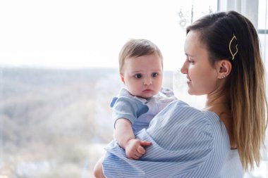 Genç bir anne kucağında küçük bir çocuk, pencerenin yanında kalıyor.