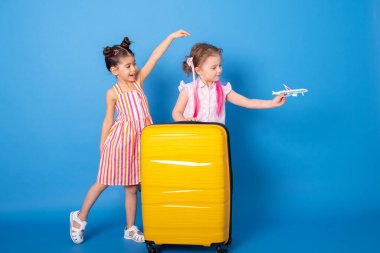 Mutlu küçük kızlar, renkli kıyafetler içinde oyuncak uçakları ve mavi arka planda sarı bavulları olan kızlar.