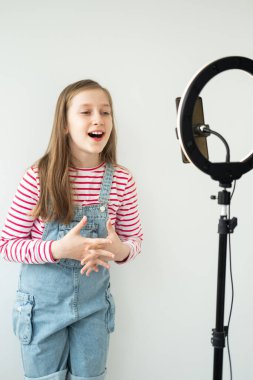 Genç kız sosyal medya blogcusu tripodda halka ışığıyla akıllı telefona bakarak video kaydediyor.