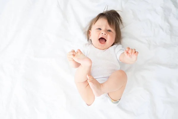 funny baby boy in white bodysuit smiling and lying on a white bedding at home. top view