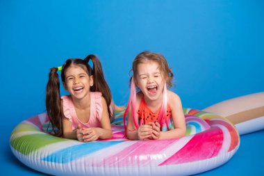 İki mutlu kız mayo giymiş renkli şişme yatakta uzanmış mavi arka planda lolipop.