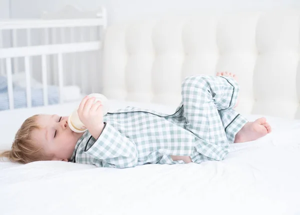 Малыш в пижаме лежит на кровати и пьет молоко из бутылочек. Детская жизнь на открытом воздухе — стоковое фото