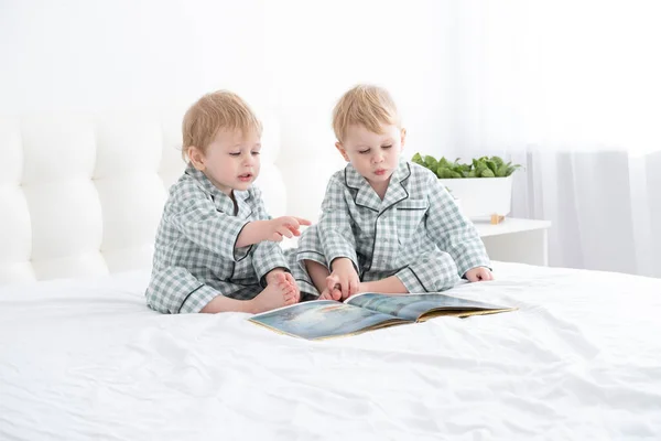 Два малыша-близнеца в пижаме читают книгу, сидя на белых кроватях на кровати. — стоковое фото
