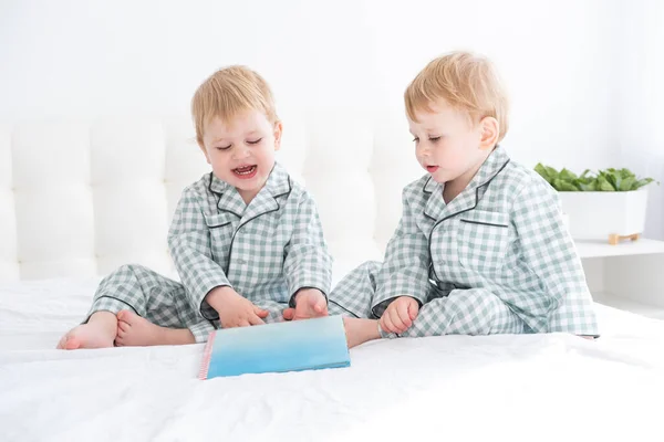 Два счастливых маленьких мальчика-близнеца в пижаме читают книгу в постели родителей — стоковое фото