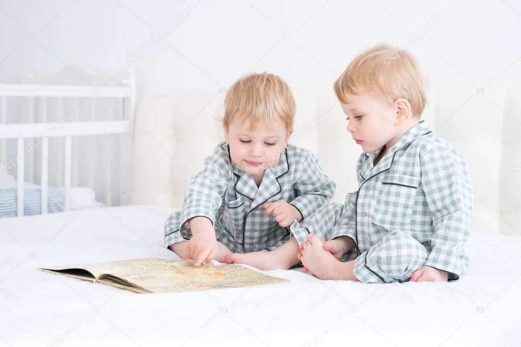 Two toddler baby twin boys in pajamas reading book sitting on white bedding on bed.