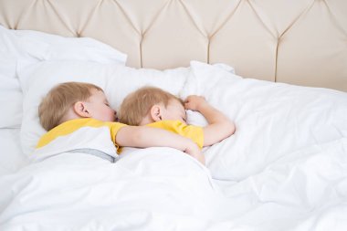 İki sarışın erkek ikiz, beyaz yatak üzerinde sarılıp uyuyorlar. .