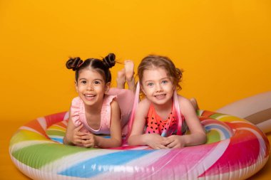 Renkli şişme yatakta uzanan mayolu iki mutlu kız sarı arka planda lolipop.