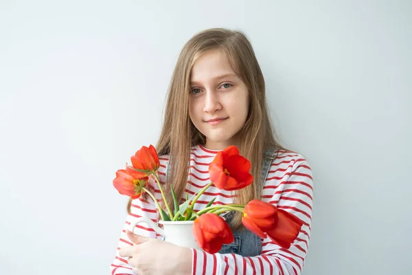 Счастливая девочка-подросток с длинными волосами, держащая вазу с красными тюльпанами, стоящими напротив белой стены — стоковое фото