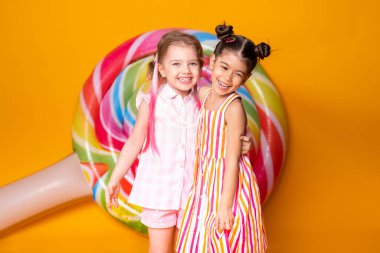 Renkli elbiseli iki mutlu kız gülüyor sarı arka planda lolipopla sarılıp eğleniyorlar.