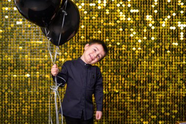 Arkaplanda altın rengi pullar ve kovalarla siyah balonlar tutan çocuk.