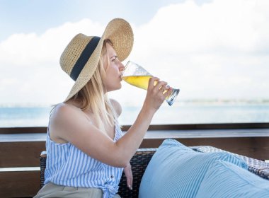 Hasır şapkalı çekici genç sarışın kadın terasta dinleniyor, sahildeki barda bira içiyor. 