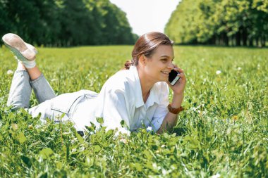 Beyaz gömlekli güzel genç kız telefonda konuşuyor parkta çimlerin üzerinde yatıyor..