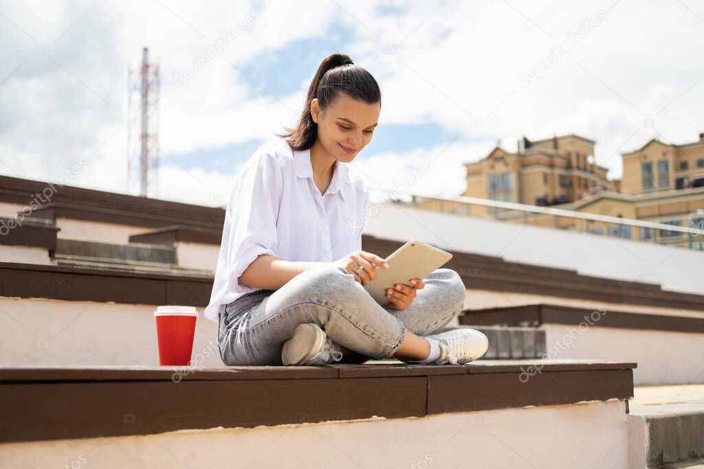 Happy young women holding tablet holding with coffee paper cup enjoying sunny day sitting in city