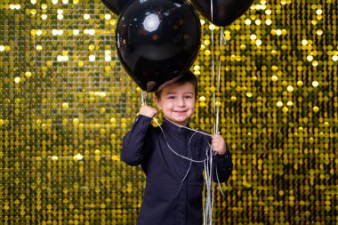 Arkaplanda altın rengi pullar ve kovalarla siyah balonlar tutan çocuk..