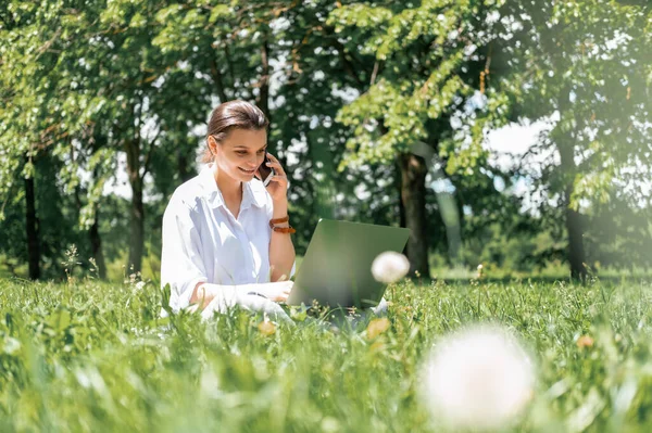 Привлекательные деловые женщины в белой рубашке разговаривают по телефону с помощью ноутбука, сидя на траве в парке. — стоковое фото