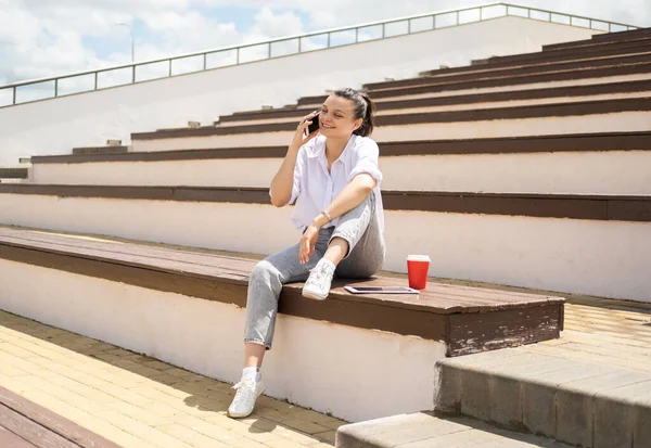Mutlu genç kız elinde kahve fincanıyla telefonda konuşuyor güneşli günün tadını çıkarıyor amfitiyatroda oturuyor. — Stok fotoğraf