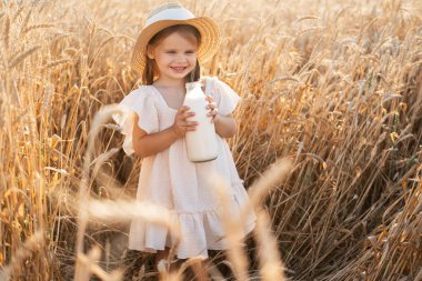 Hasır şapkalı ve bej kumaşlı çocuk gün batımında buğday tarlasında süt savaşı veriyor..