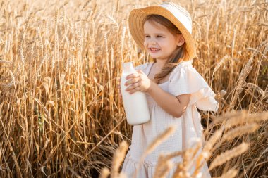 Hasır şapkalı ve bej kumaşlı çocuk gün batımında buğday tarlasında süt savaşı veriyor..