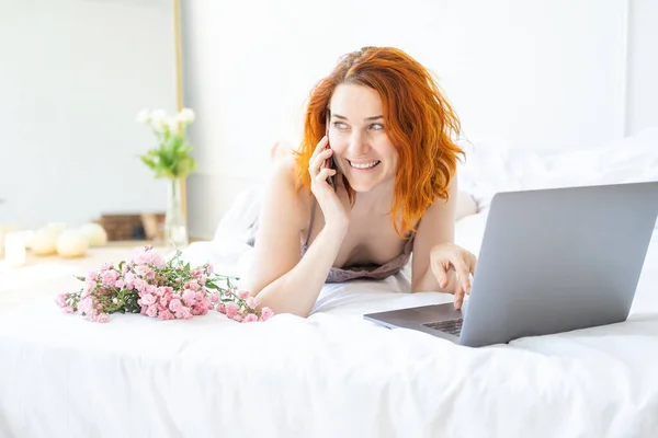 Atractiva mujer jengibre de mediana edad en pijama acostado en la cama utilizando el teléfono y el ordenador portátil —  Fotos de Stock