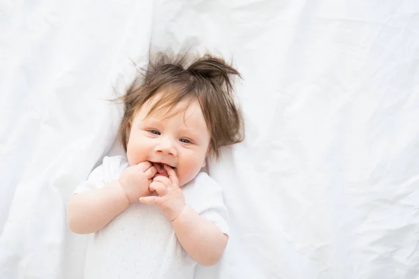 Retrato de menina engraçada com o dedo em uma boca sorrindo e deitado em uma cama branca em casa. — Fotografia de Stock