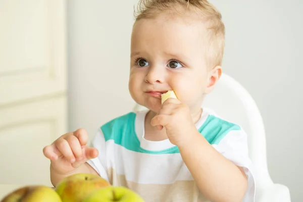 可愛い男の子が子供の椅子に座って白いキッチンでリンゴを食べる. — ストック写真