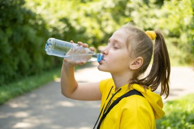 Sarı elbiseli genç kız parkta plastik şişeden su içiyor.