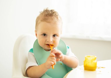 Önlüklü komik çocuk çocuk çocuk koltuğunda oturmuş kaşıkla sebze püresi yiyor. Çocuk kendi başına yemek yemeyi öğreniyor..