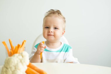 Bebek çocuk Childs sandalyesinde oturup beyaz arka planda havuç dilimleri yiyor.