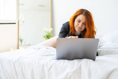 Çekici, güler yüzlü, orta yaşlı bir kadın parlak odada dizüstü bilgisayar kullanarak yatakta yatıyor..