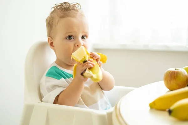 子供の椅子に座って白いキッチンでバナナを食べる面白い赤ちゃん — ストック写真