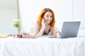 atraktivní zázvor žena středního věku v pyžamu ležící v posteli pomocí telefonu a notebooku.
