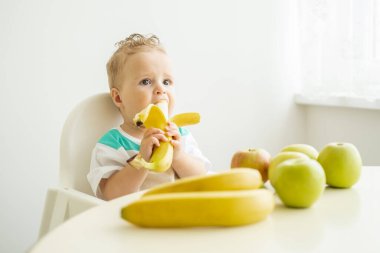 Çocuk sandalyesinde oturan komik çocuk beyaz mutfakta muz yiyor.