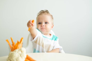 Bebek çocuk Childs sandalyesinde oturup beyaz arka planda havuç dilimleri yiyor..