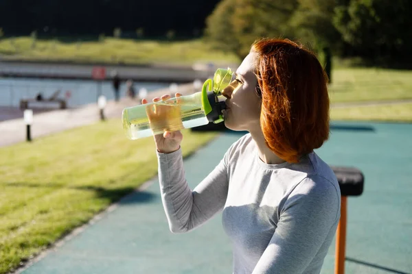 秋のスポーツグラウンドで晴れた日に仕事をしながら水を飲むスポーツ服の生姜女に合う — ストック写真