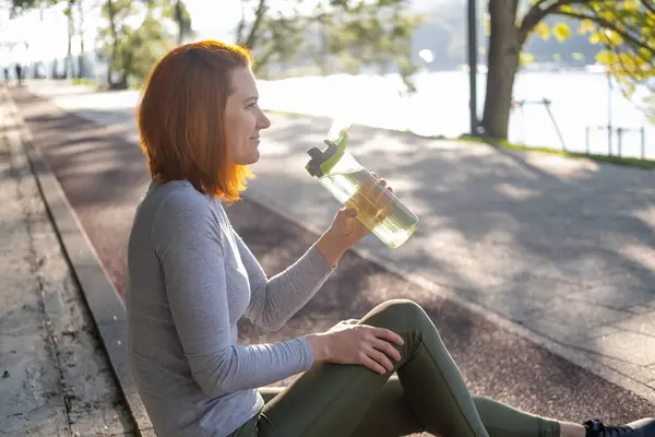 Štíhlá zázvorová běžkyně ve sportovním oblečení sedící v parku a pijící vodu. Zdravý fitness životní styl — Stock fotografie