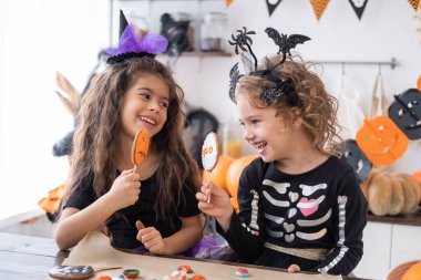 Cadı kostümü giymiş iki farklı çocuk mutfakta eğleniyor, kurabiye yiyor, Cadılar Bayramı 'nı kutluyor..