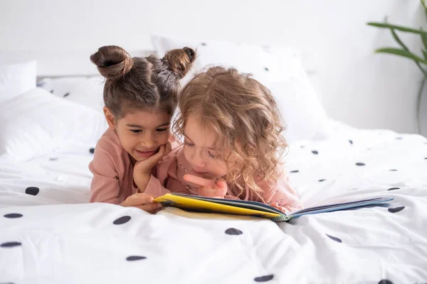 Две Разноплановые Девочки Сестры Подруги Пижаме Читают Книгу Лежа Белом — стоковое фото