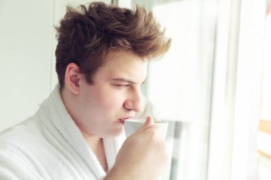 Genç beyaz adam pencerenin yanında kahve içiyor. O bir kedi tutuyor. Sabah erken, gün ışığı. 