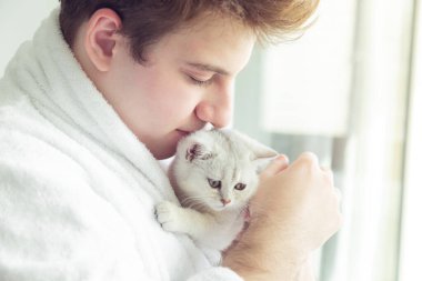 Bornozlu genç beyaz bir adam beyaz bir kedi yavrusu tutuyor. Sabahın erken saatlerinde pencerenin yanında durur.. 