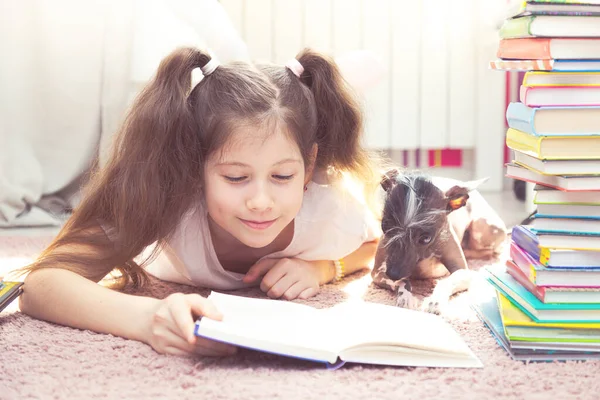 小女孩和一只狗在地板上玩耍 他们周围有许多书 女孩试图看书 但被狗打扰了 — 图库照片